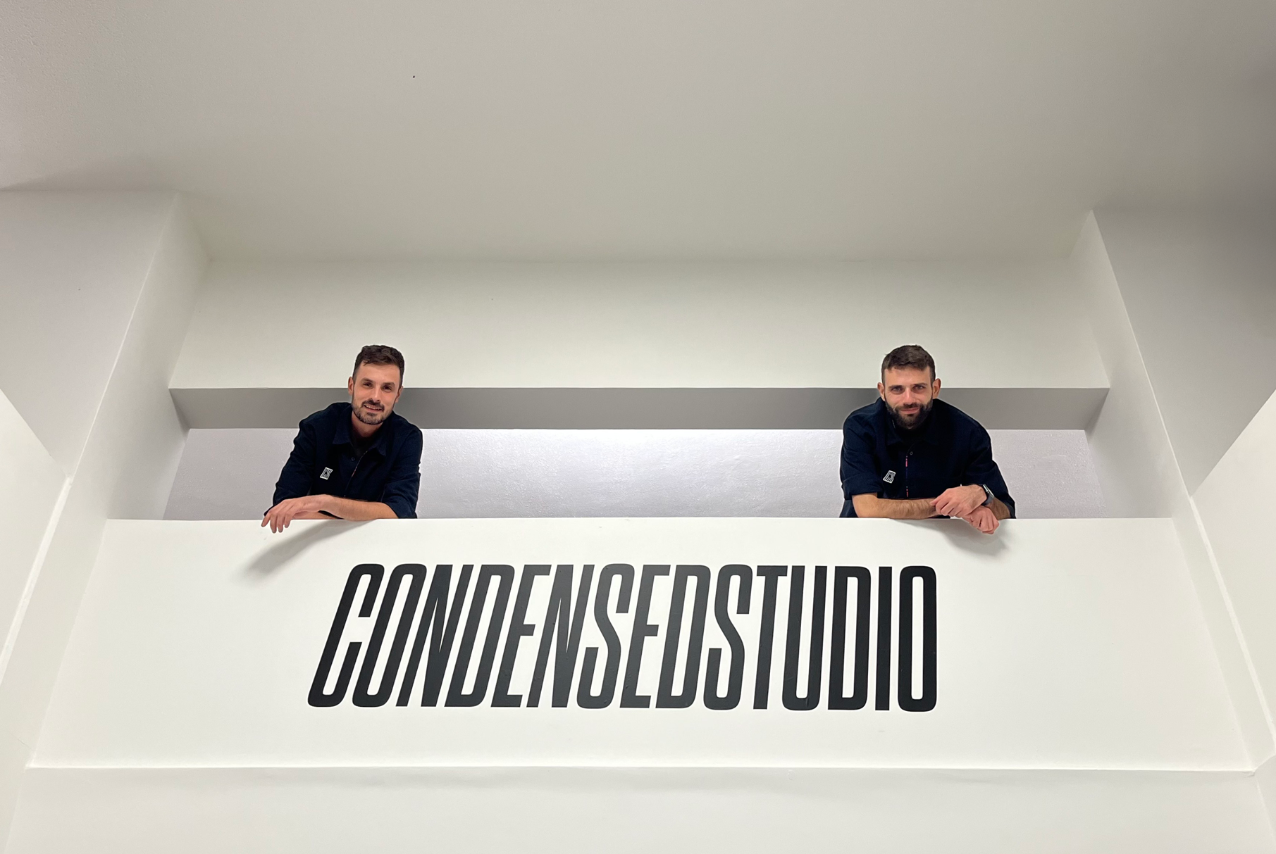 Marco e Stefano appoggiati su una balaustra bianca con il logo 'CONDENSED STUDIO' in caratteri neri allungati. Lo sfondo è minimalista con pareti bianche e un design moderno.