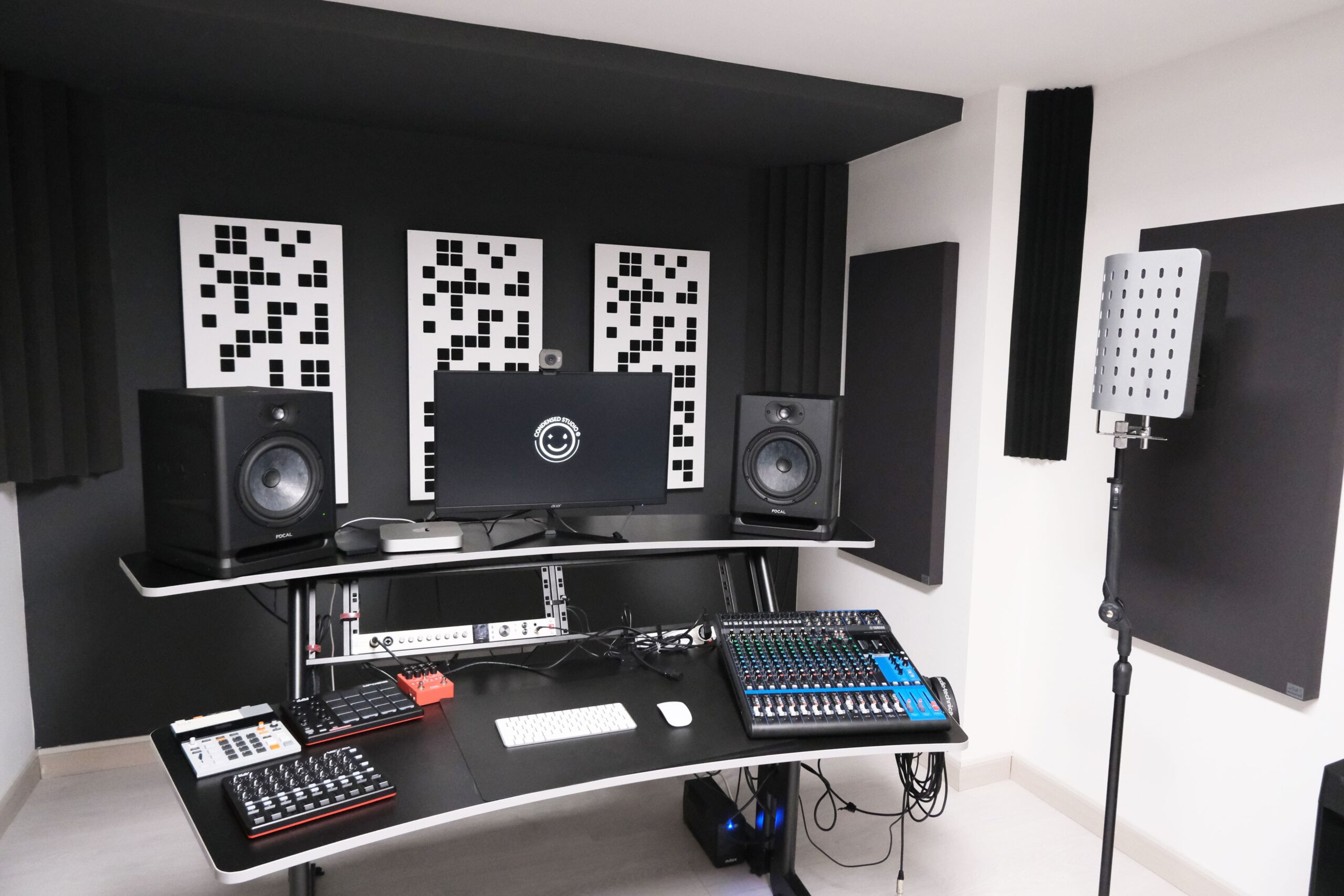 Studio di registrazione professionale con pareti insonorizzate e pannelli acustici decorativi. Al centro, una scrivania con monitor, interfaccia audio, mixer, tastiera MIDI e altre attrezzature per la produzione musicale. Due casse monitor Focal sono posizionate ai lati dello schermo. A destra, un microfono con filtro antiriflesso su un'asta. L'ambiente è moderno e ben organizzato.