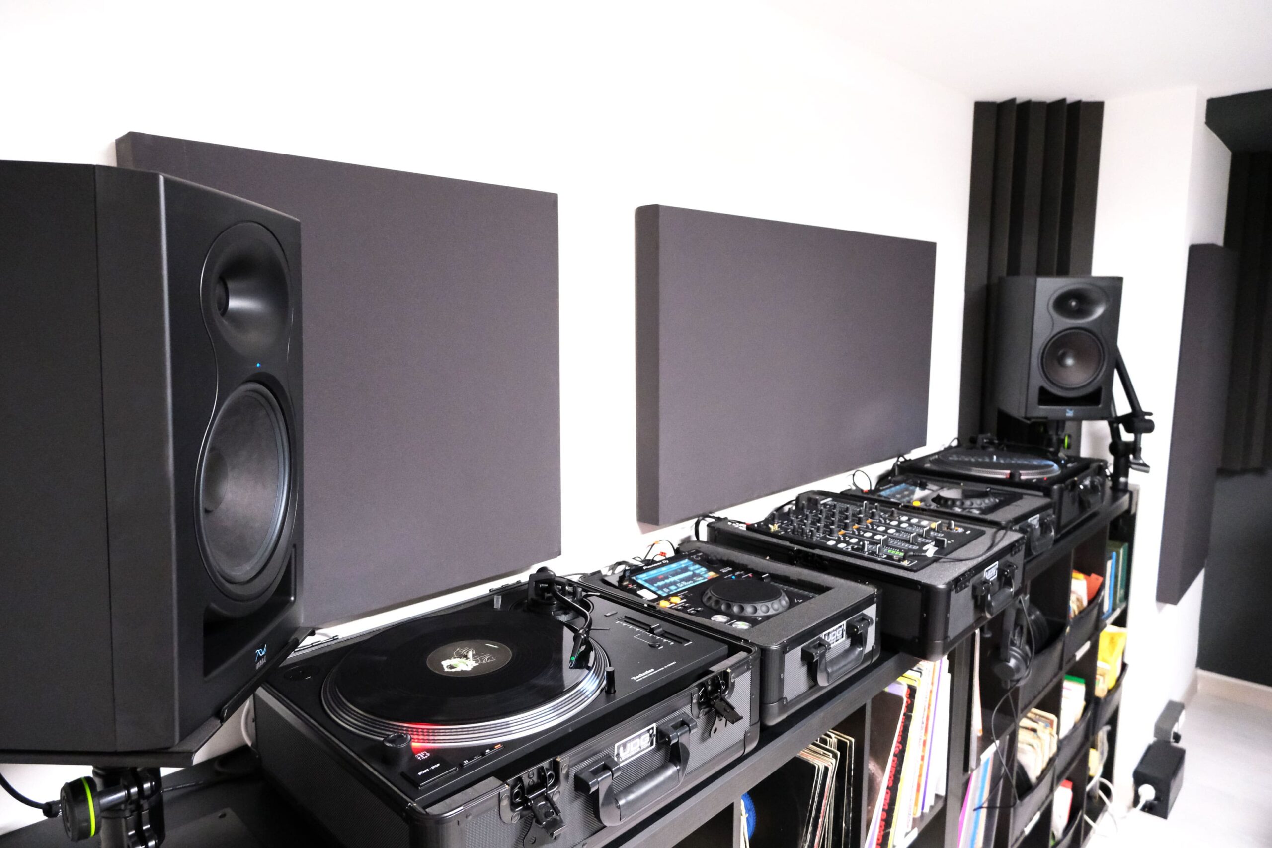 Setup DJ professionale con due giradischi Technics, mixer, lettore CDJ e casse monitor su una scrivania attrezzata. I dispositivi sono posizionati su flight case per la protezione e la portabilità. Sullo sfondo, pannelli fonoassorbenti per un'acustica ottimale. Sotto la postazione, una libreria piena di vinili e accessori per DJing