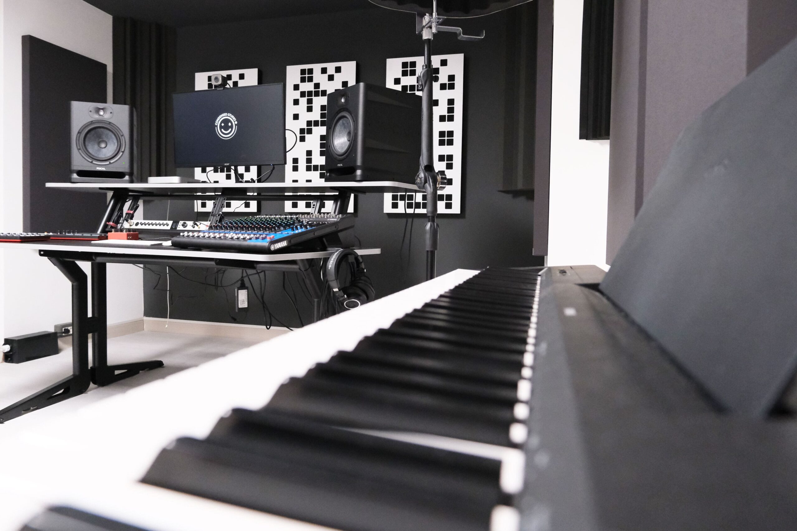 Condensed Studio, sala di registrazione moderno con pianoforte digitale in primo piano, console di missaggio Yamaha, monitor audio Focal e pannelli acustici geometrici sulle pareti.